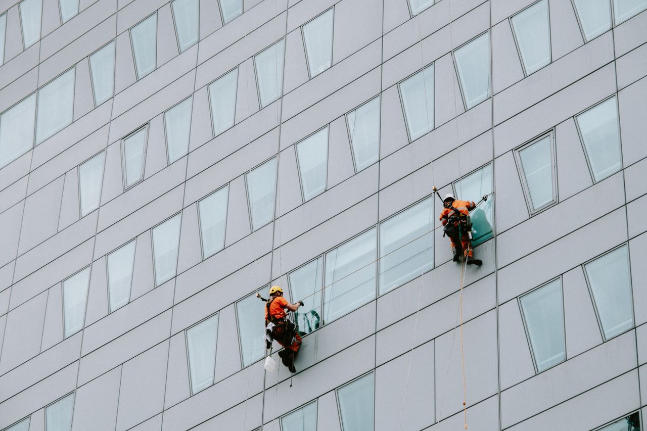 services-03 Des laveurs de vitres en hauteur, équipés de protections, nettoient la façade d’un gratte-ciel.
